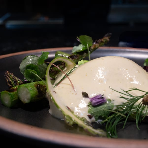 Ember's dish with asparagus, creamy sauce, fresh herbs, and purple flowers on a dark plate.